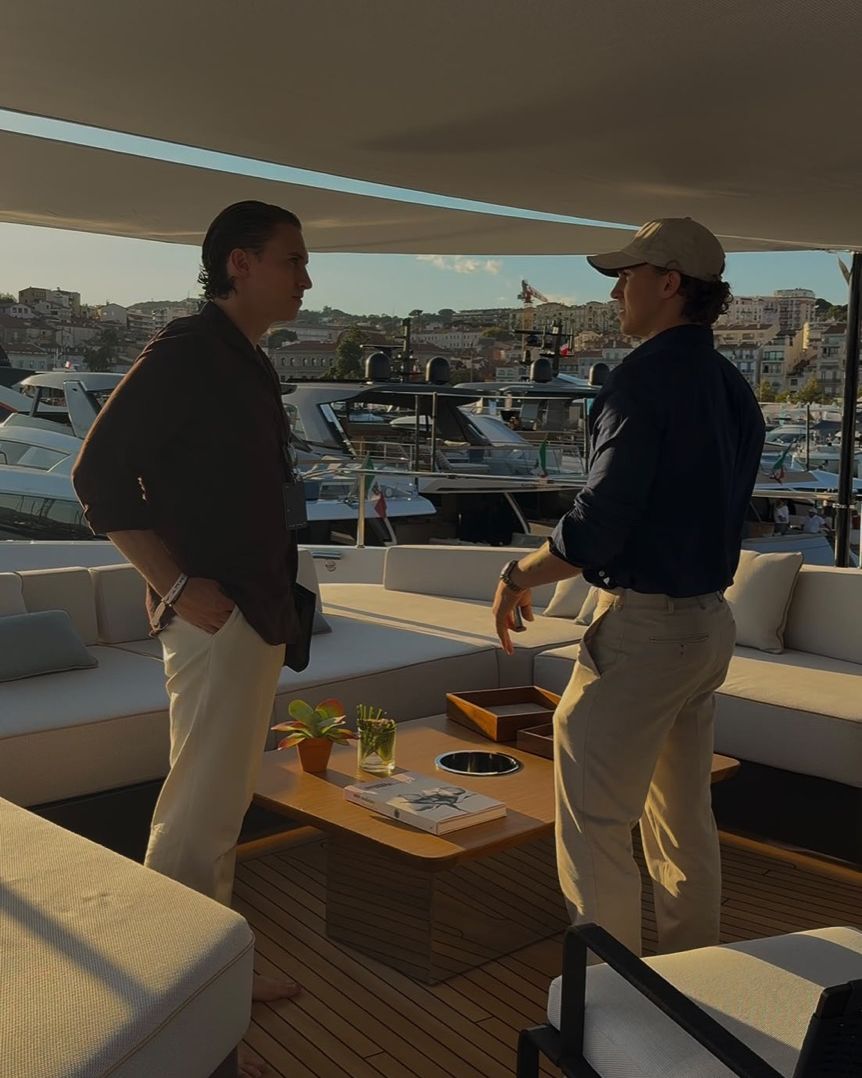 Two men in conversation on the shaded deck of a yacht, the harbour of Cannes with more yachts at the Yachting Festival behind them