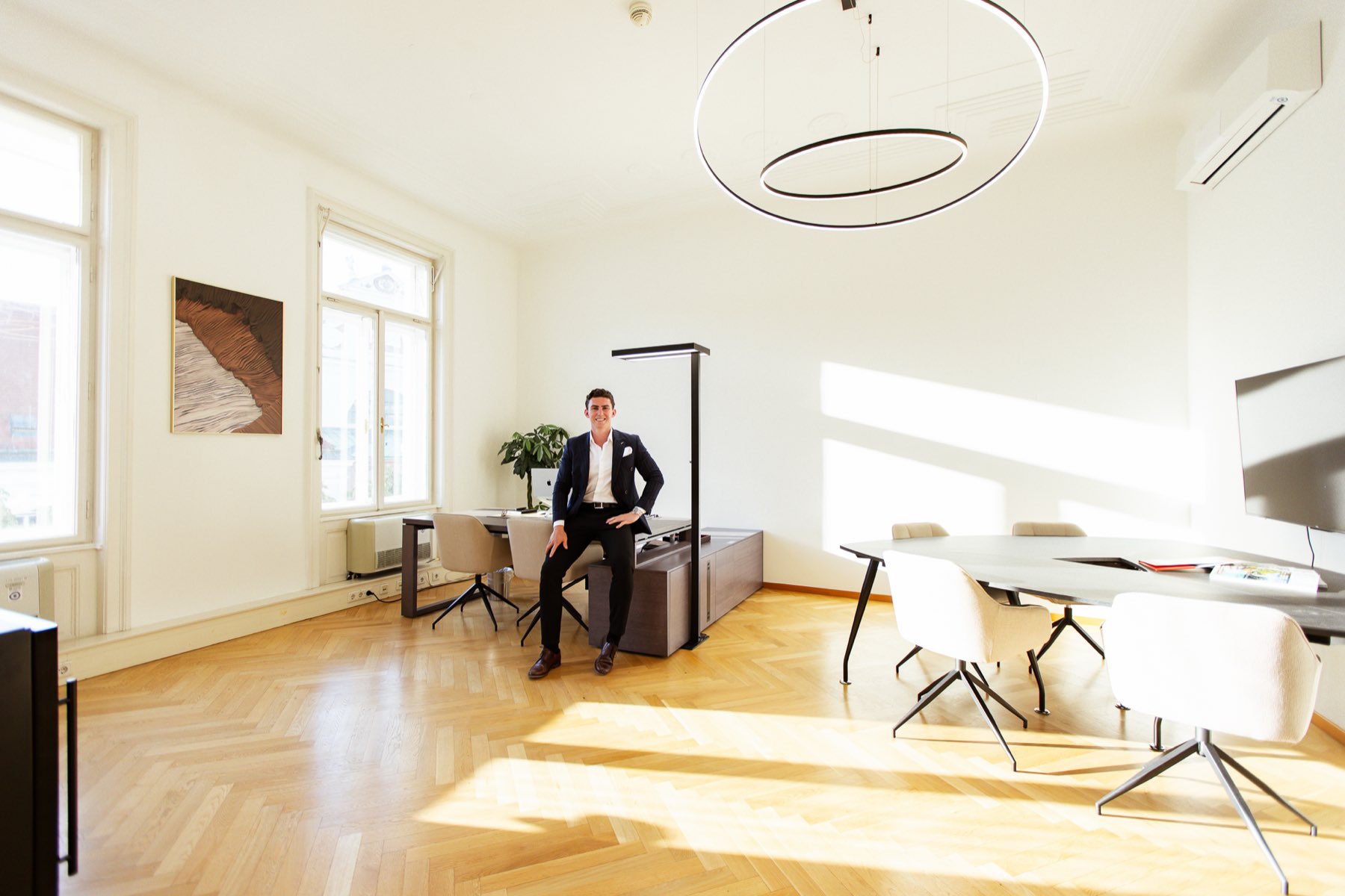 The NIC office in Vienna 1010: a sunlit period room with herringbone parquet, a large round pendant lamp and a meeting table, one of the founders in a dark suit sitting on the sideboard
